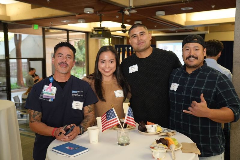 Four members of the Bruin veteran community smile together at a table decorated with small American flags during UCLA's annual Veterans Day reception at the James West Alumni Center.
