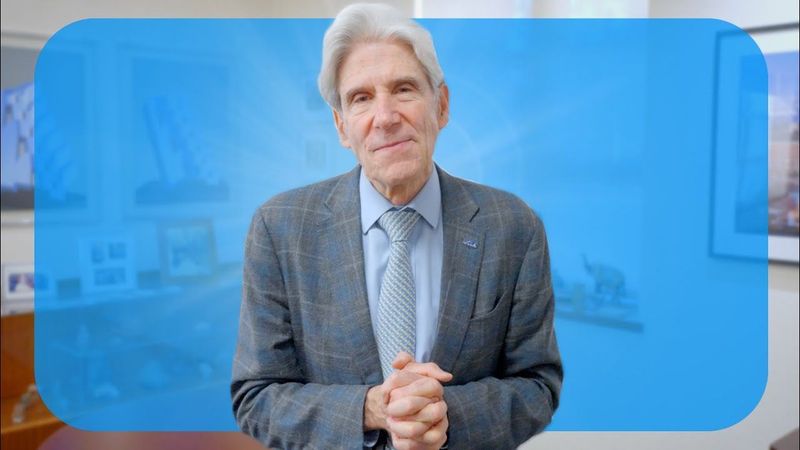 UCLA Chancellor Julio Frenk in a gray blazer smiling with his hands clasped in front of a blue background