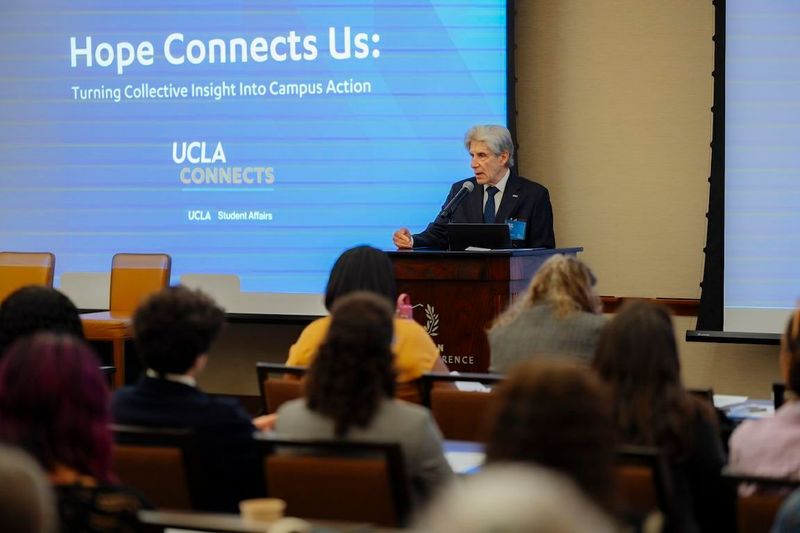 UCLA Chancellor Julio Frenk stands at a wooden podium speaking into a microphone before an audience of attendees seated at tables in a conference room. Behind him, a large blue projection screen displays the text "Hope Connects Us."