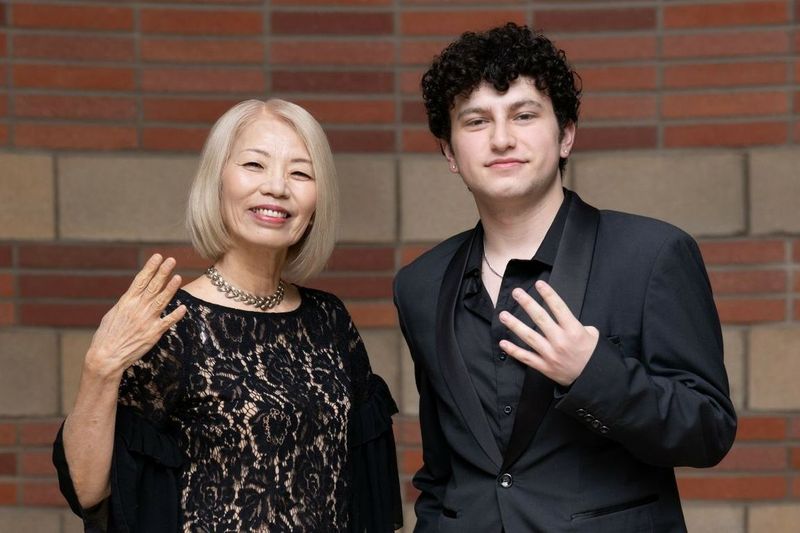 Mayu Sasame and August Maturo stand against brick wall, each smiling and holding up four fingers to signifies the UCLA eight-clap cheer