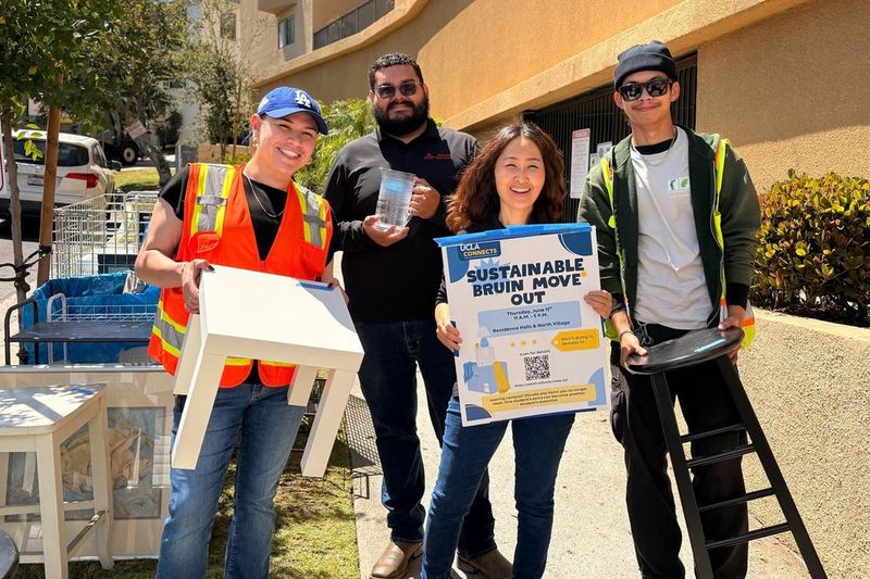 Four volunteers pose outdoors near donated furniture items, including a white table and bar stool, holding a UCLA Connects Sustainable Bruin Move Out event sign.