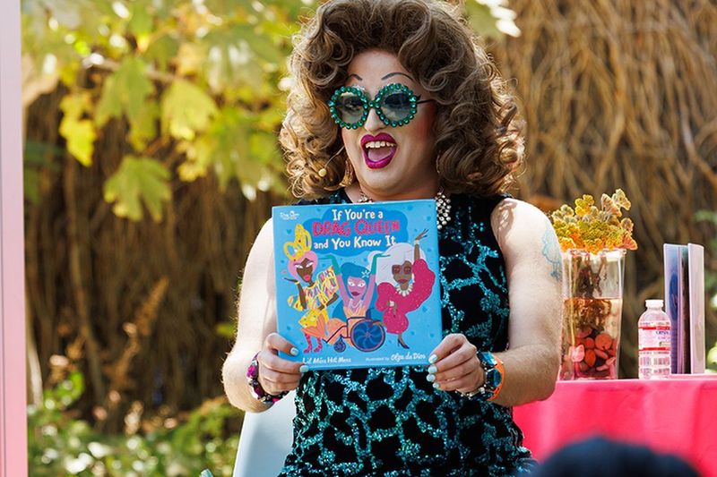 Drag performer Lil Miss Hot Mess sits outdoors reading the children’s book If You’re a Drag Queen and You Know It to an audience. Wearing oversized jeweled sunglasses and a sparkling blue-and-black outfit, they hold the book open while smiling animatedly.