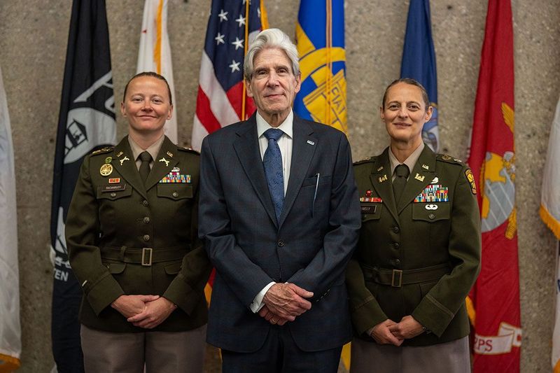 Chancellor Julio Frenk, wearing a dark navy suit and a blue-patterned tie, stands in the center between two U.S. Army officers in olive-green dress uniforms adorned with medals and ribbons.